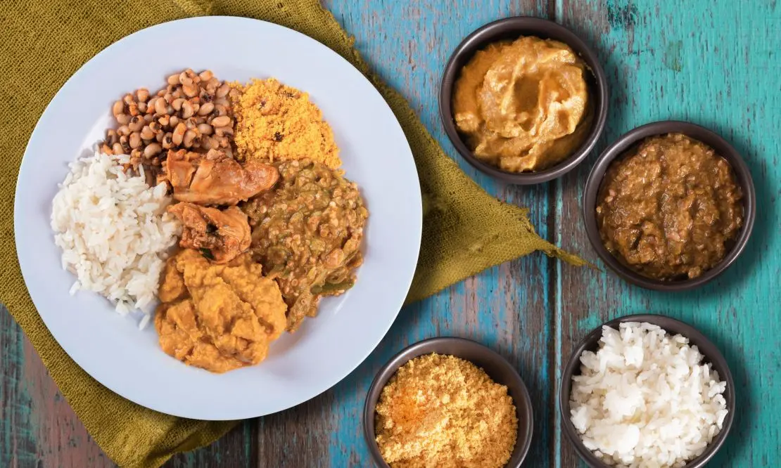 visão de cima de um prato sobre uma mesa de madeira. O prato tem comidas típicas do Brasil como arroz, feijão e caruru