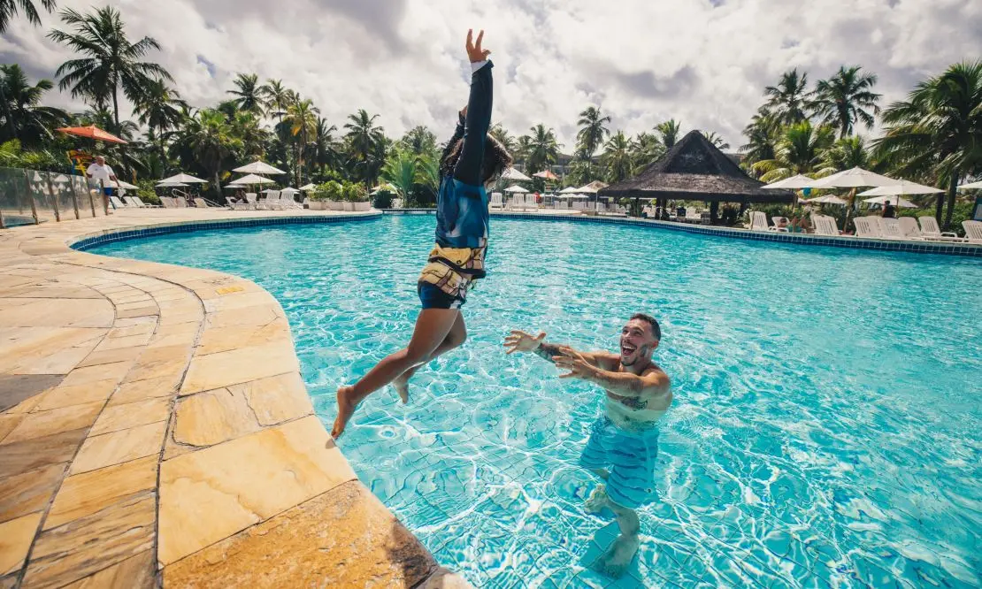 Criança saltando na piscina e um homem esperando na água para segurá-la