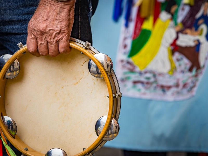 Parte das mãos de um homem que segura um pandeiro, e uma bandeira colorida da Folia de Reis ao fundo