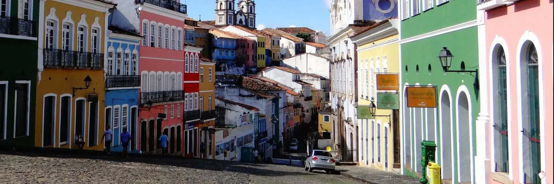 Imagem de uma rua numa das cidades turísticas da Bahia. Nela, é possível ver casas coloridas e alguns carros.