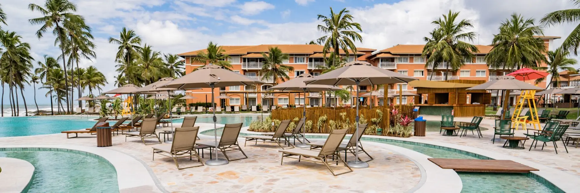 Imagem da piscina de um resort all inclusive. Na beirada dela, é possível ver algumas mesas com guarda-sol.