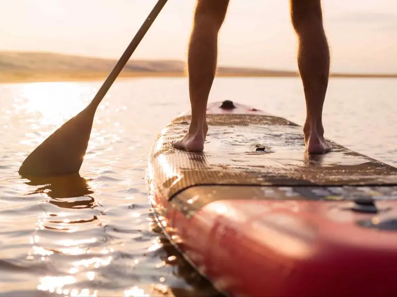 Parte das pernas de uma pessoa sobre uma prancha de SUP, remando em direção ao sol que está se pondo