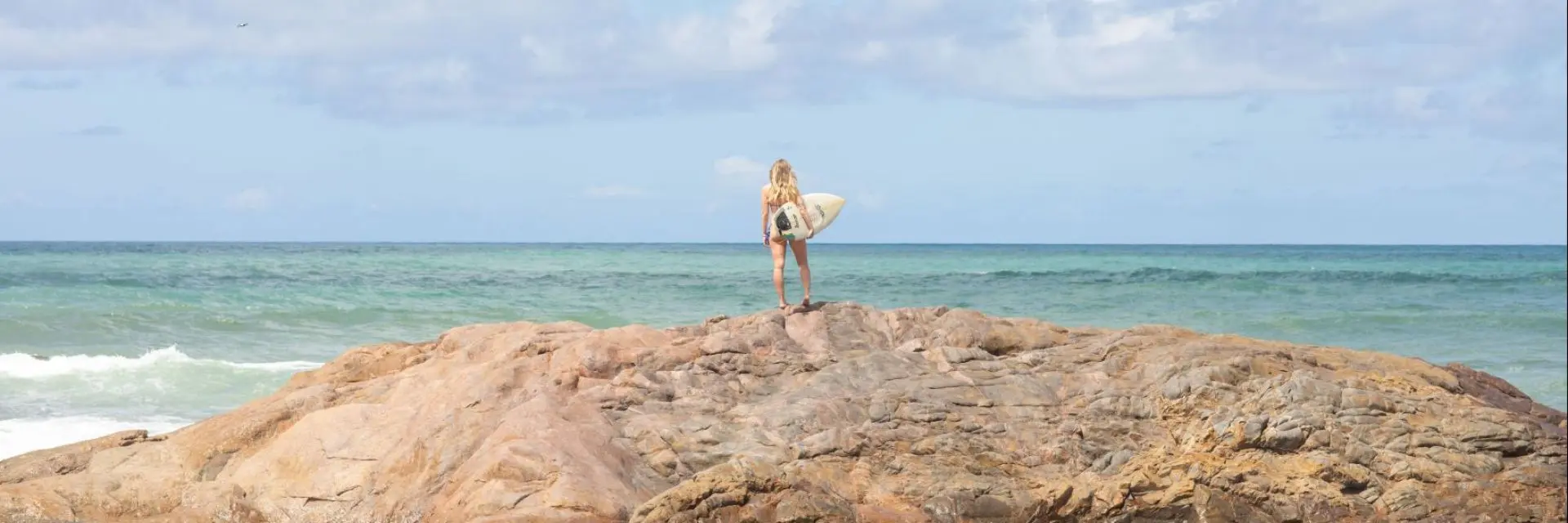 Mulher parada em cima de um conjunto de rochas com uma prancha de surf na mão em frente ao mar e céu azul.