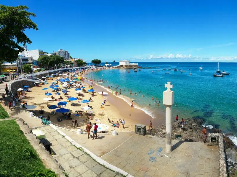 Praia de Porto da Barra e cruz de fundação da região. A areia da praia está chega de pessoas com guarda-sóis coloridos 