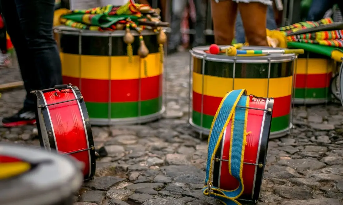 Instrumentos de percussão coloridos no chão enquanto algumas parte das pernas de algumas pessoas estão paradas ao lado dos instrumentos