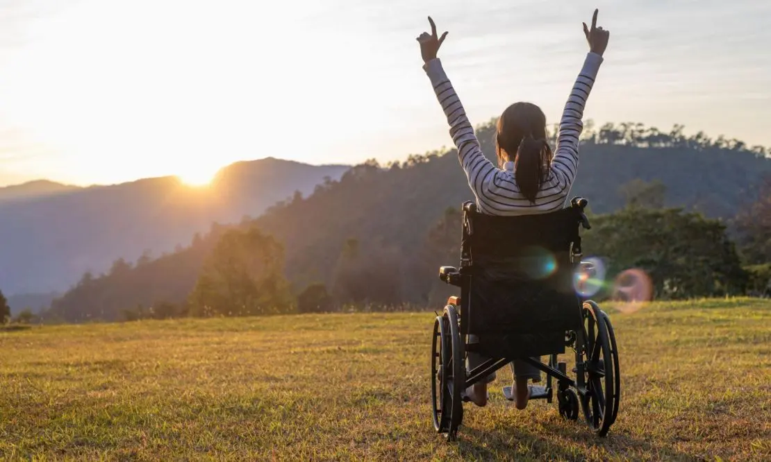Mulher de costas em uma cadeira de rodas, com os braços levantados admirando o nascer do sol