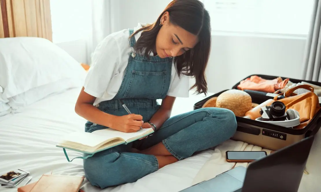 Mulher sentada na cama planejando uma viagem com um caderno no colo e uma mala aberta