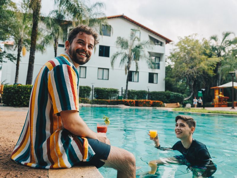 Uma pessoa sentada na beira de uma piscina no Rio Quente e segurando um coquetel, enquanto outra está dentro da piscina, também segurando uma bebida colorida