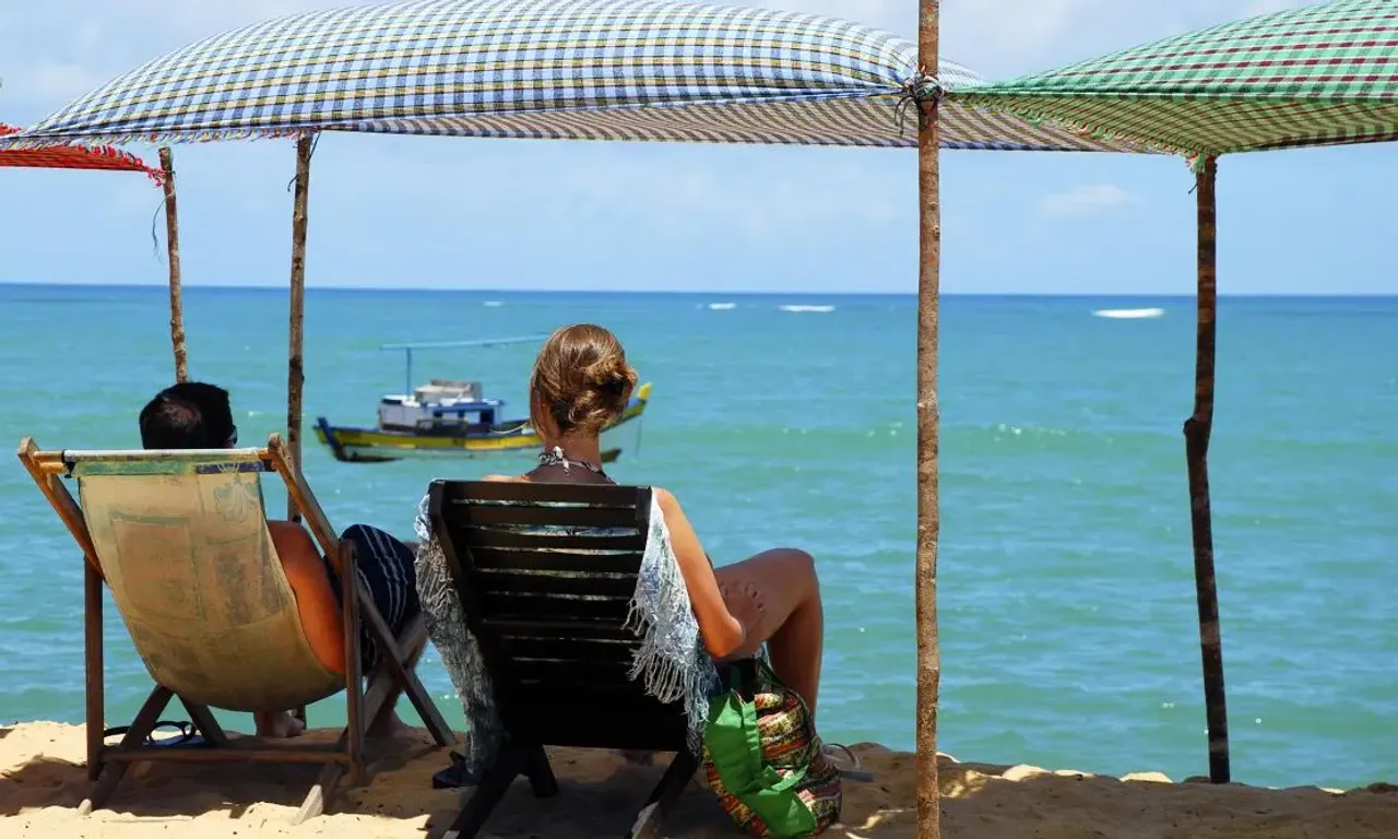 Casal sentado em cadeiras de pano sobre um guarda sol em Porto seguro