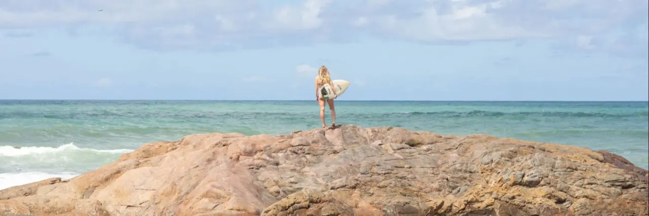 Mulher parada em cima de um conjunto de rochas com uma prancha de surf na mão em frente ao mar e céu azul.