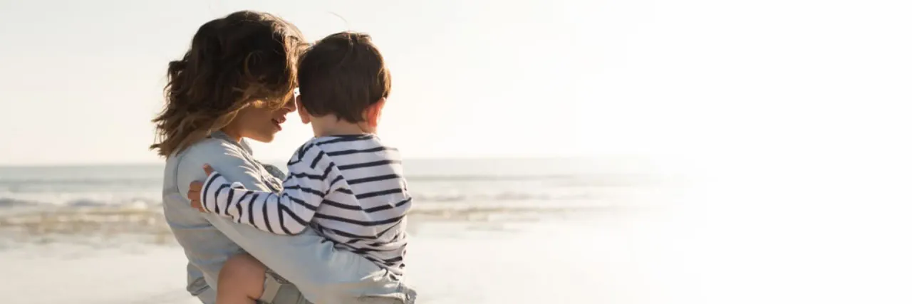Imagem de uma mulher de costas segurando um bebê. Eles estão observando a areia e o mar de uma praia 