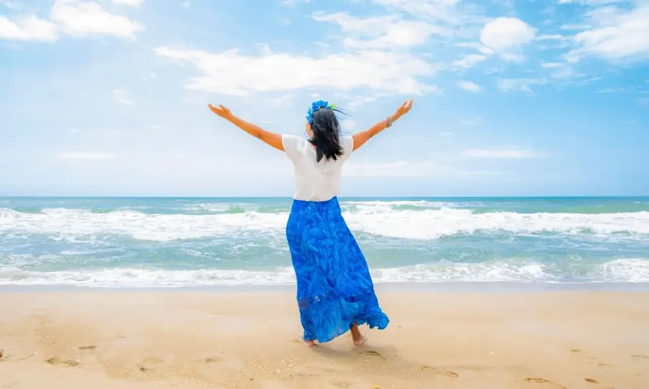 Mulher de costas usando saia longa azul e camiseta branca, olhando para o mar de braçoa abertos